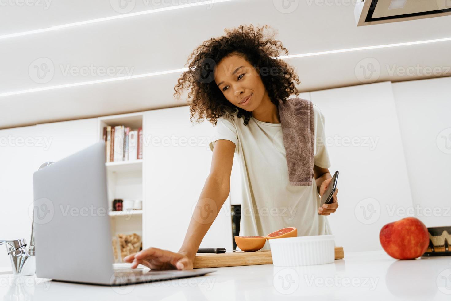 Mujer joven negra con portátil mientras prepara el desayuno en la cocina foto