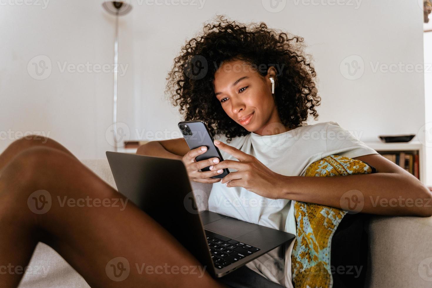 Black young woman using mobile phone and laptop while resting on sofa chair at home photo