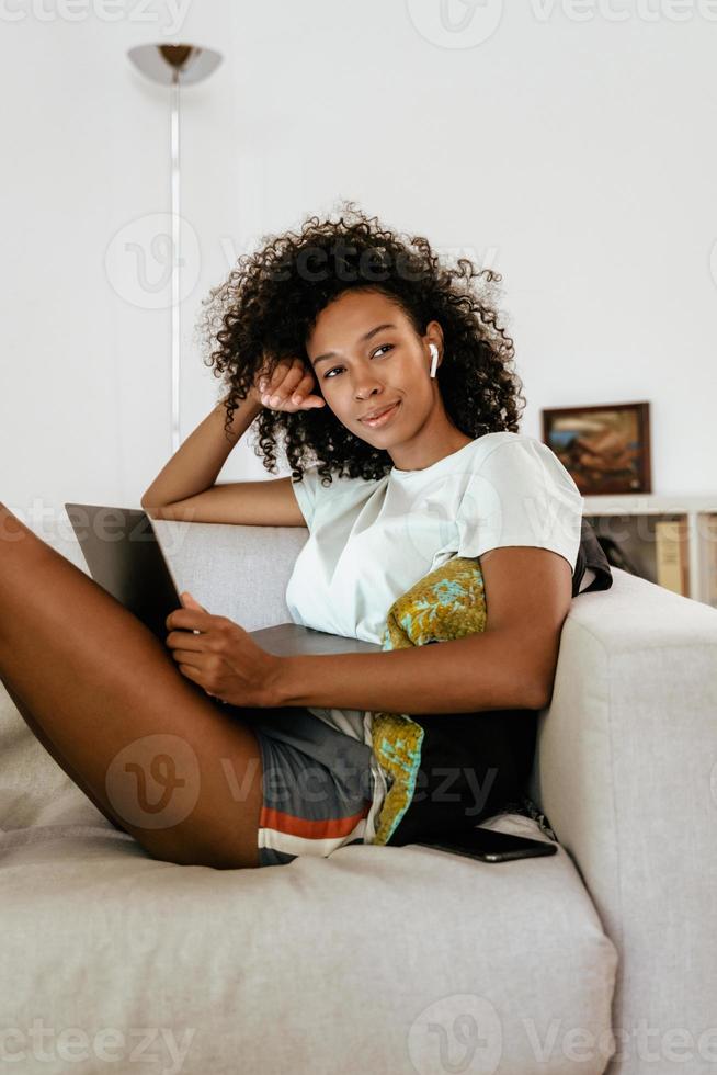 Black young woman in earphones using laptop while resting on sofa and looking at the camera photo