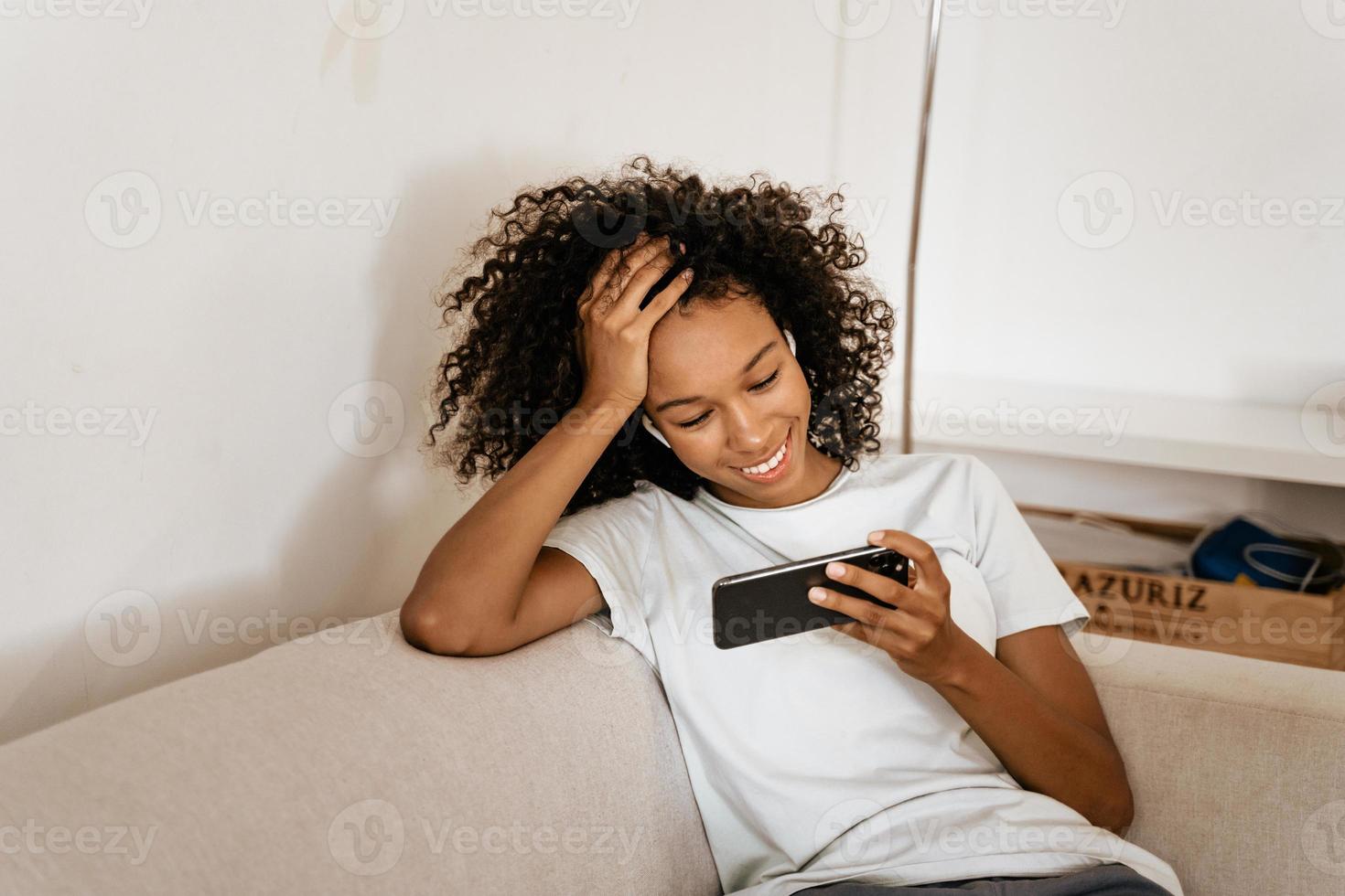 Black young woman with earphones uses mobile phone while resting on sofa photo