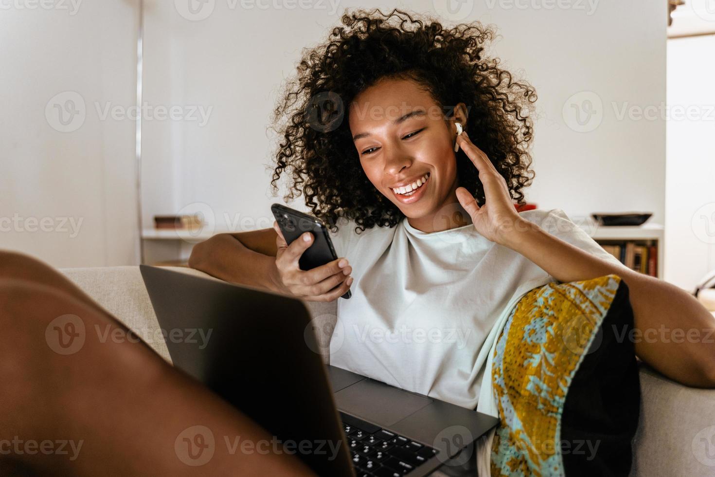 Black young woman using mobile phone with headphones and laptop while resting on sofa photo