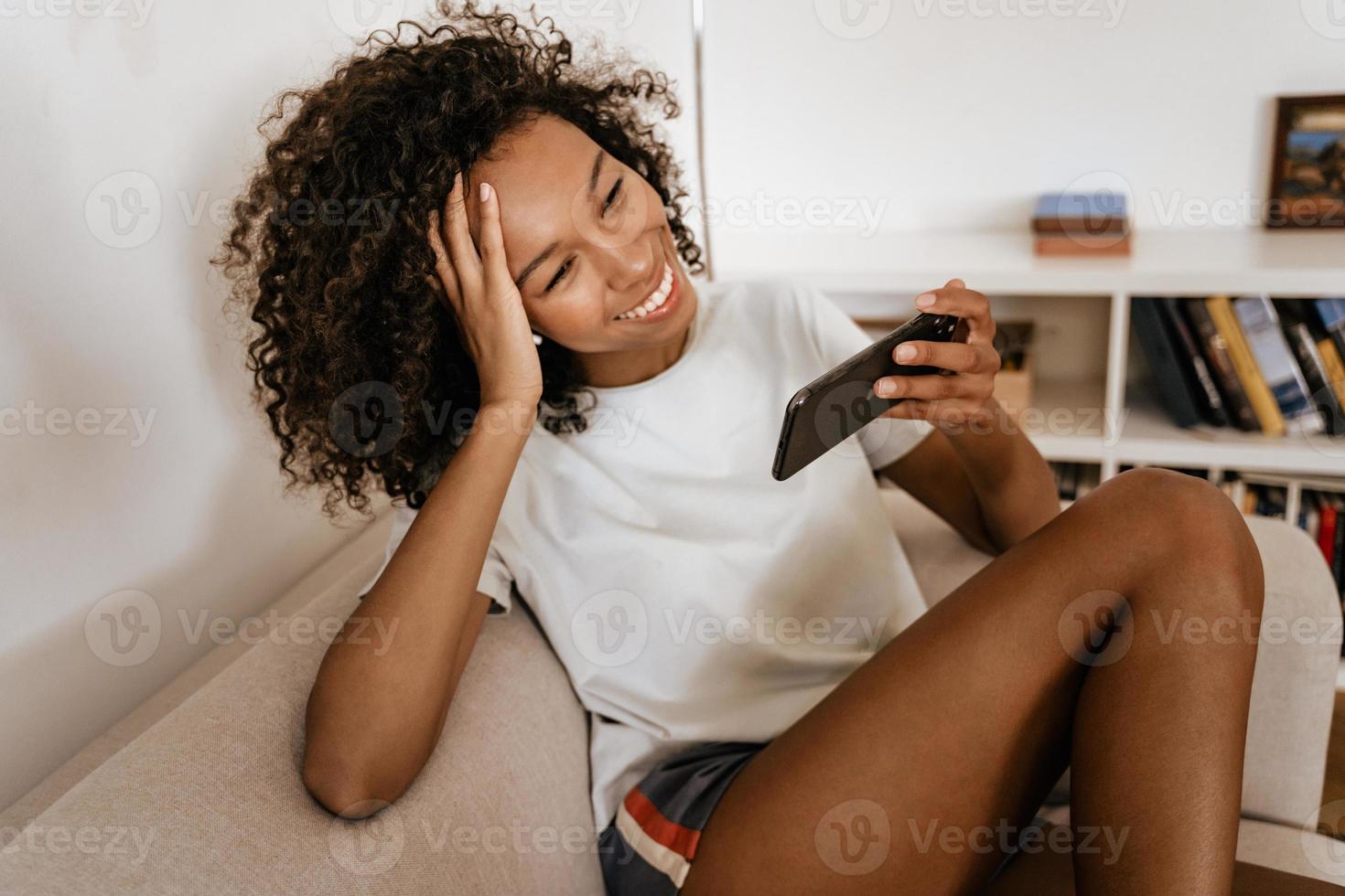 Young Black woman in earphones using mobile phone while resting on sofa photo