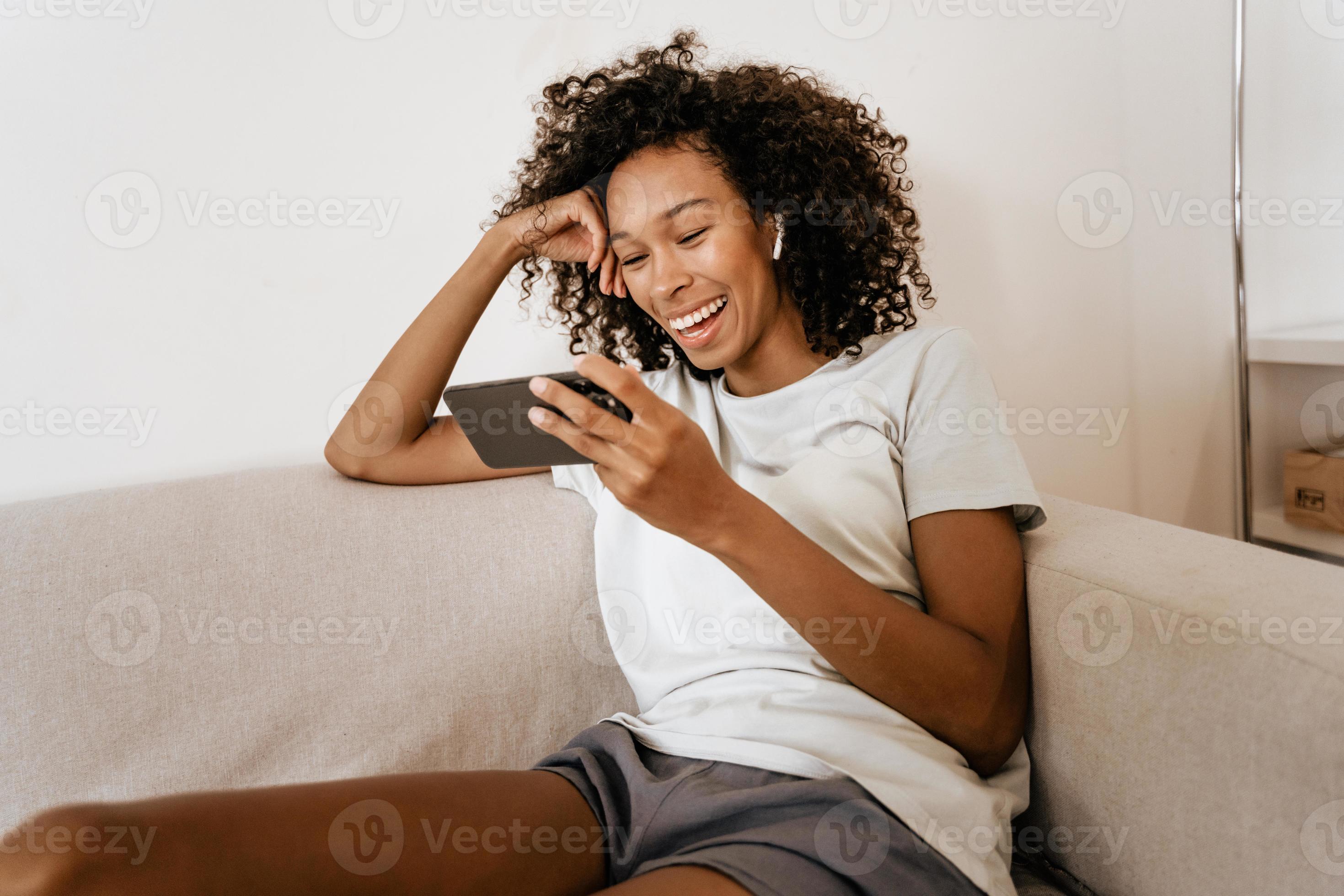 Black smiling young woman in earphones using mobile phone while laying on sofa photo