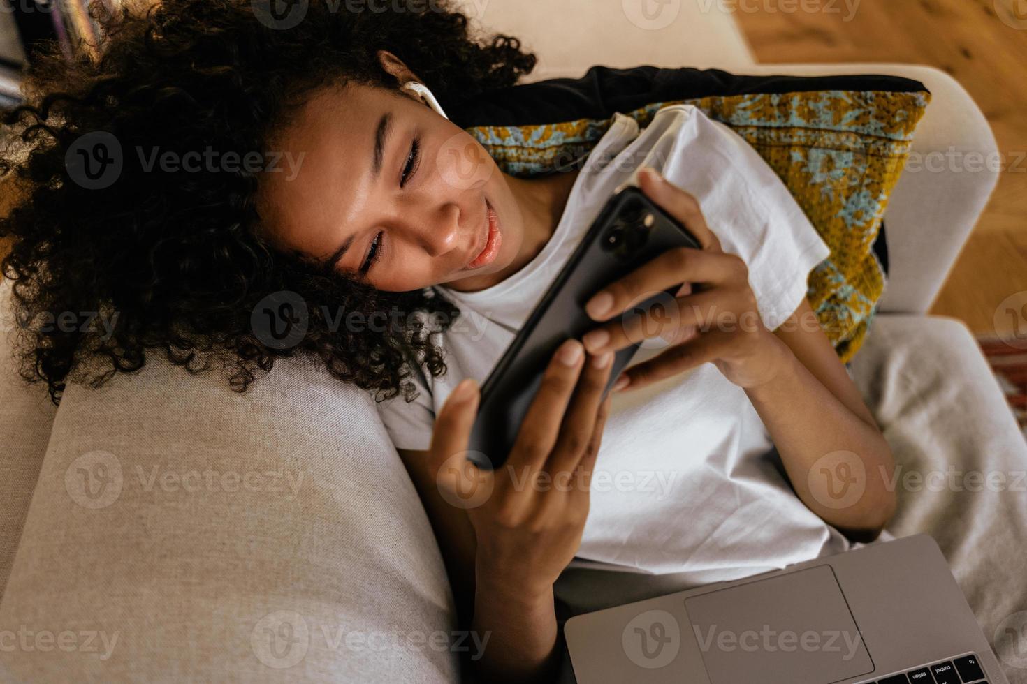 Joven negra con teléfono móvil y portátil mientras descansa en el sofá foto