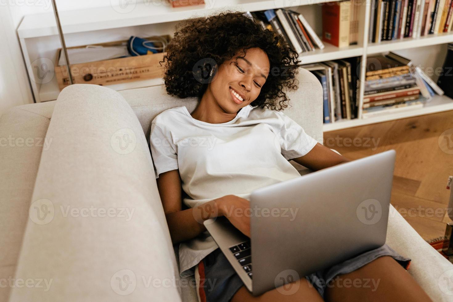 Black young woman in earphones using laptop while resting on sofa photo