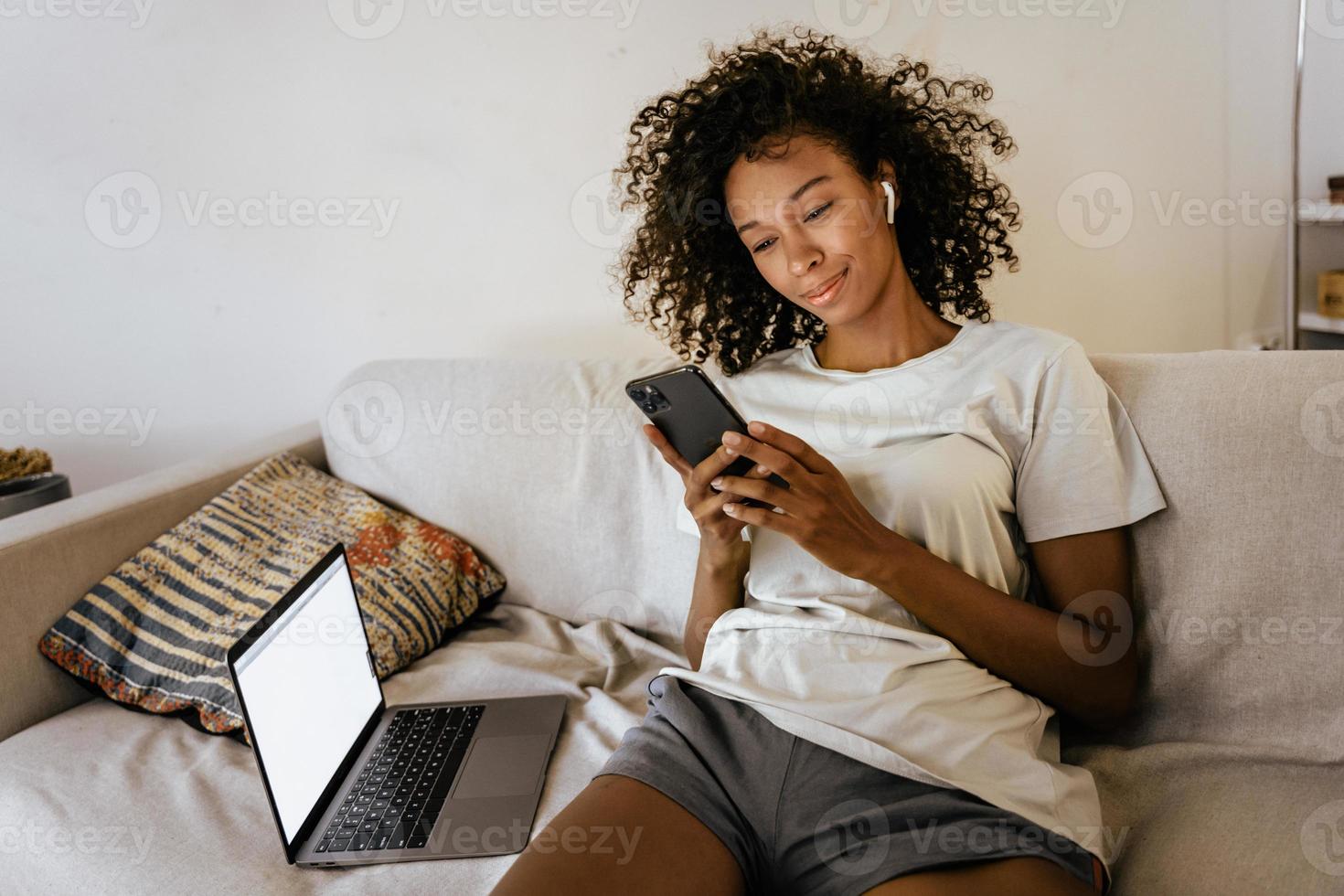 Black young woman in earphones using cellphone while resting on sofa photo
