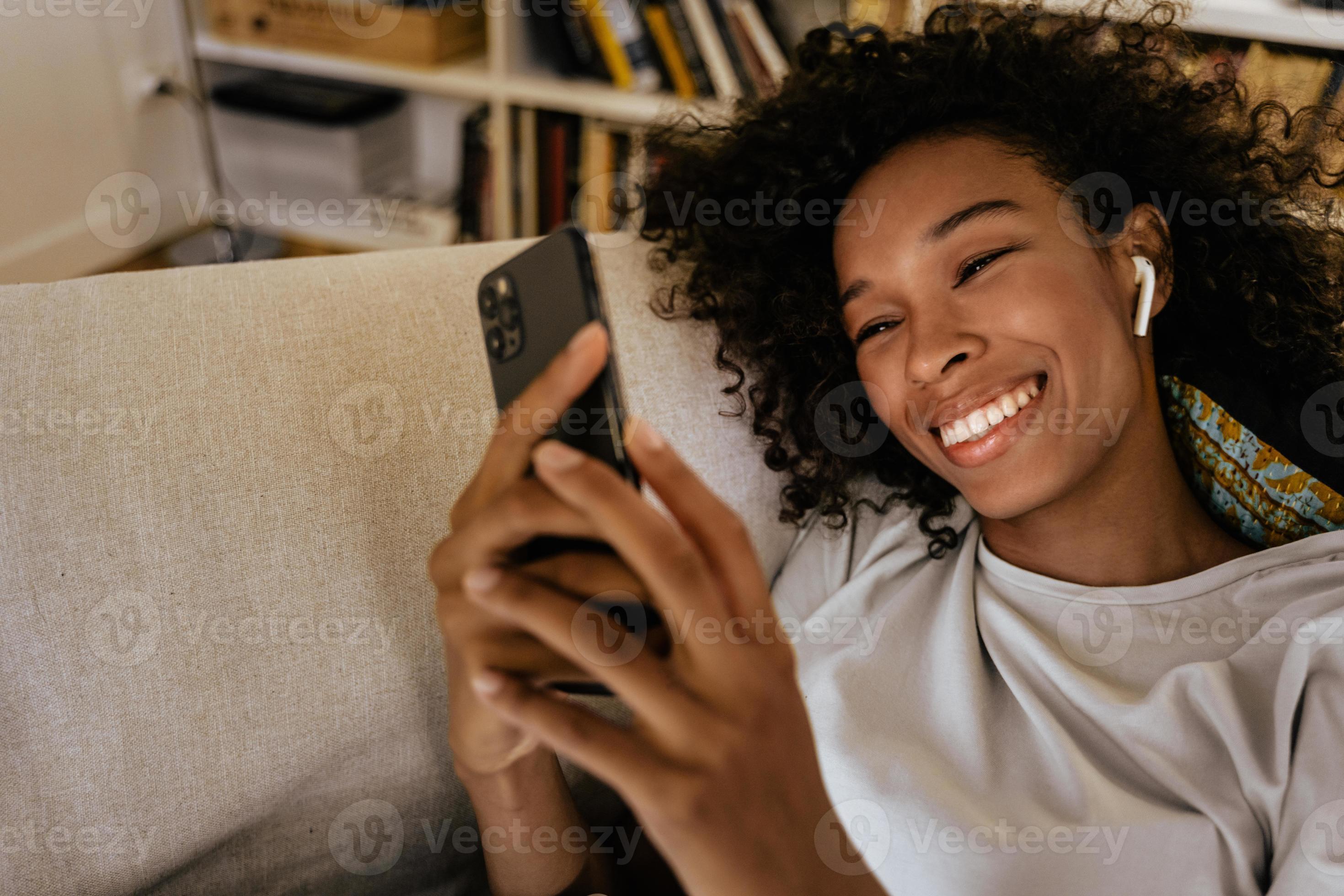 Black young woman in earphones using mobile phone while resting on sofa photo