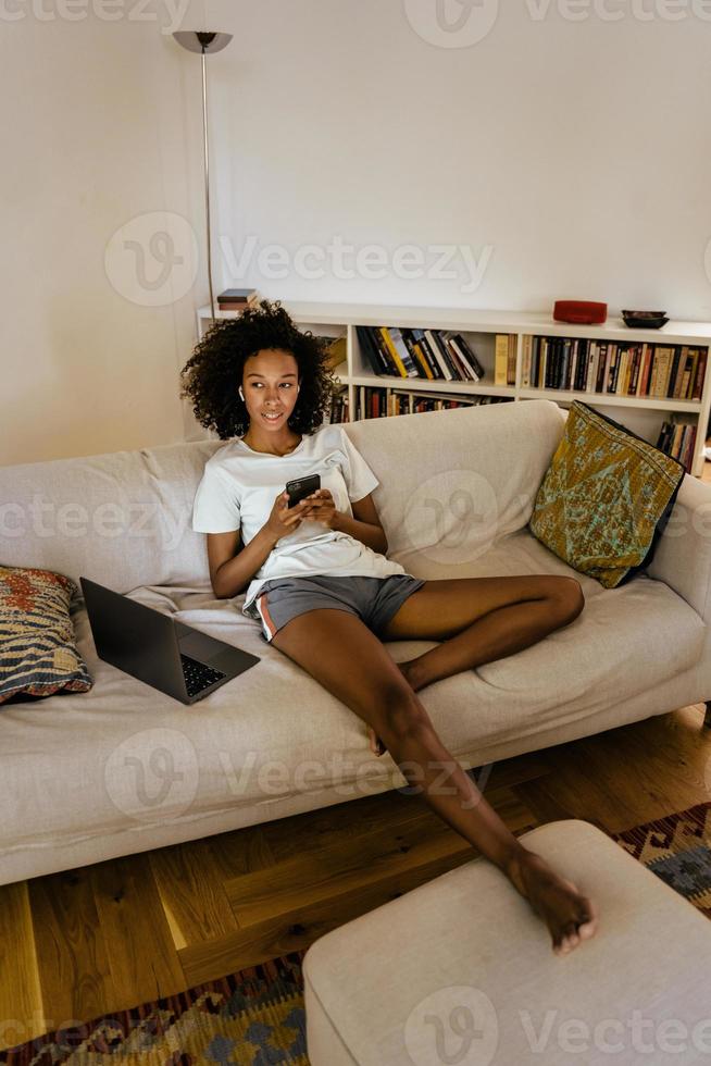 Black young woman in earphones using cellphone while resting on sofa photo