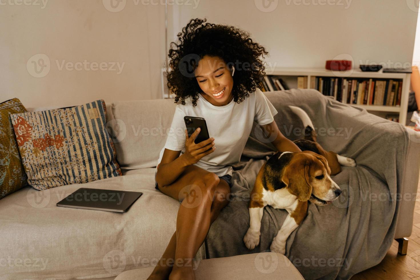 Black young woman using cellphone and stroking her dog on sofa photo