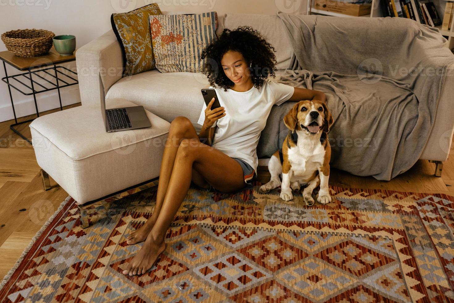 Mujer negra con teléfono celular y acariciando a su perro mientras está sentada en el piso foto