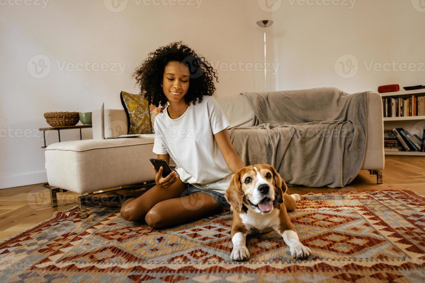 Mujer negra con teléfono celular y acariciando a su perro mientras está sentada en el piso foto