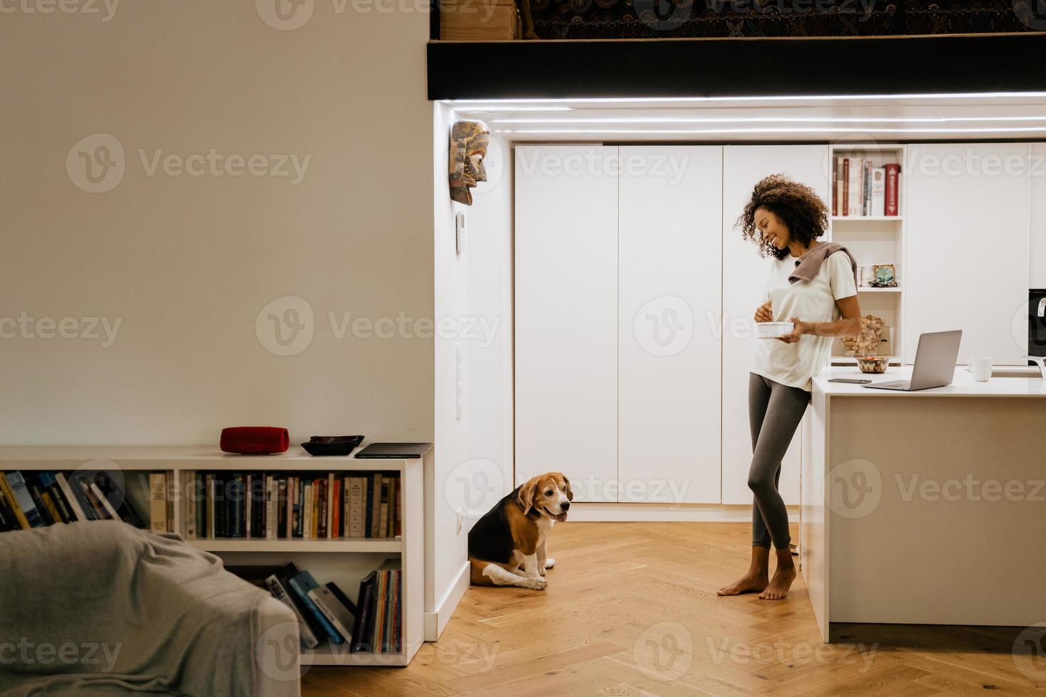 Black young woman having breakfast while looking at her dog in kitchen photo