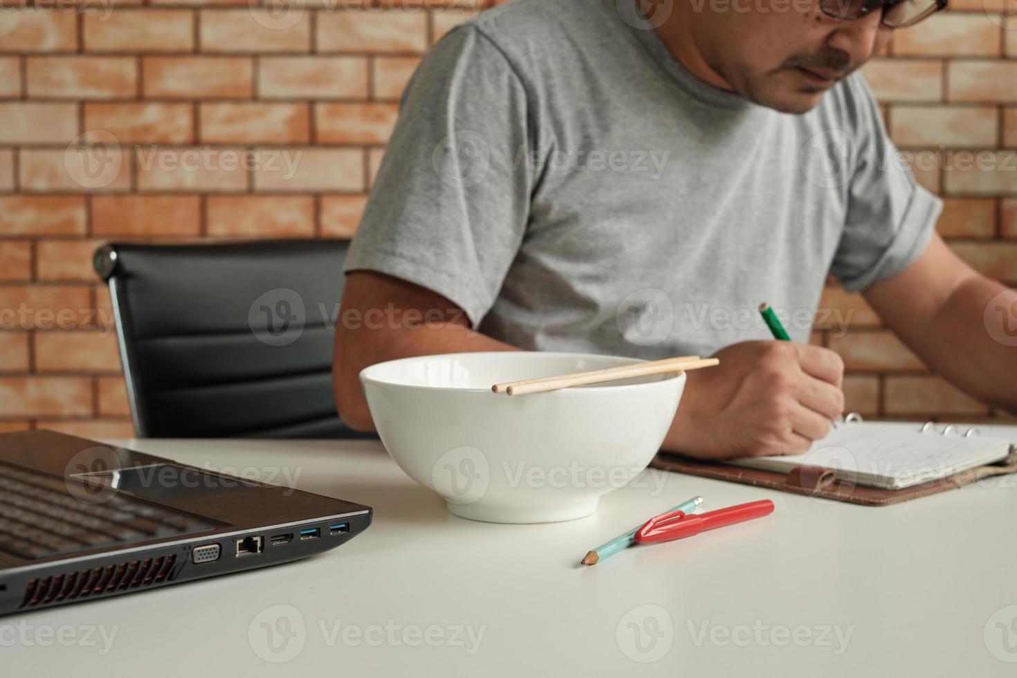 Thai male worker busy working with laptop, use chopsticks to hastily eat instant noodles during office lunch's break, because quick, tasty, and cheap. Over time Asian fast food, unhealthy lifestyle. photo