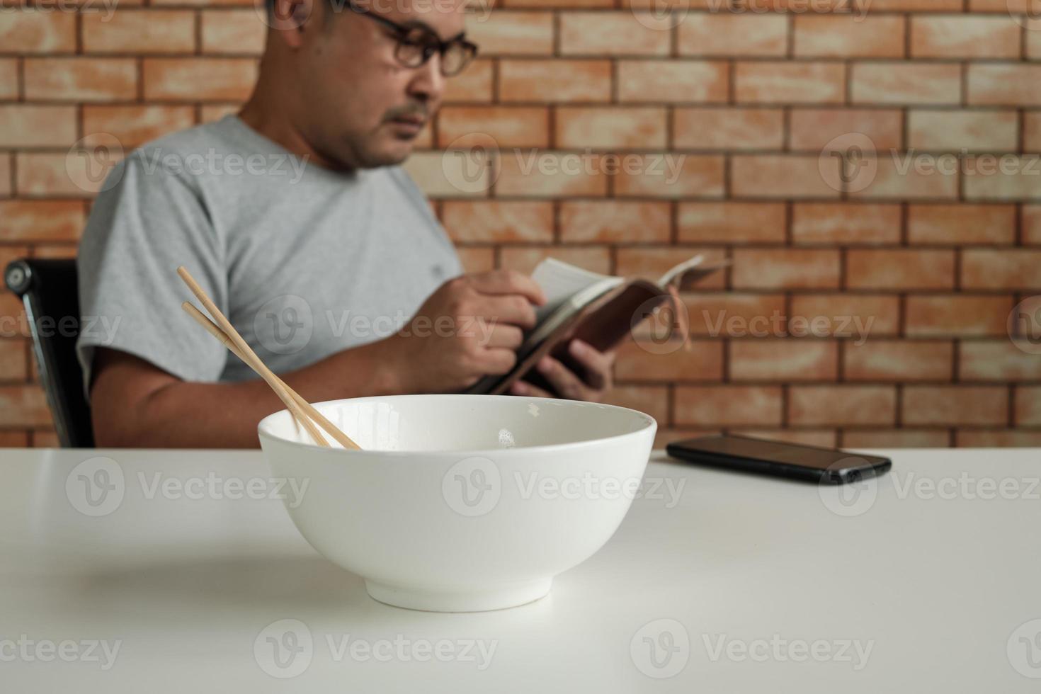 trabajador de sexo masculino asiático lee un libro de citas mientras come fideos instantáneos en un tazón blanco con palillos en la mesa en la oficina de fondo de pared de ladrillo durante un descanso a la hora del almuerzo, un estilo de vida apresuradamente insalubre. foto