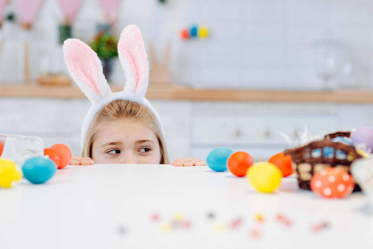 Cute little girl with white bunny ears looking at camera while peeking out from behind table with colorful Easter eggs. photo