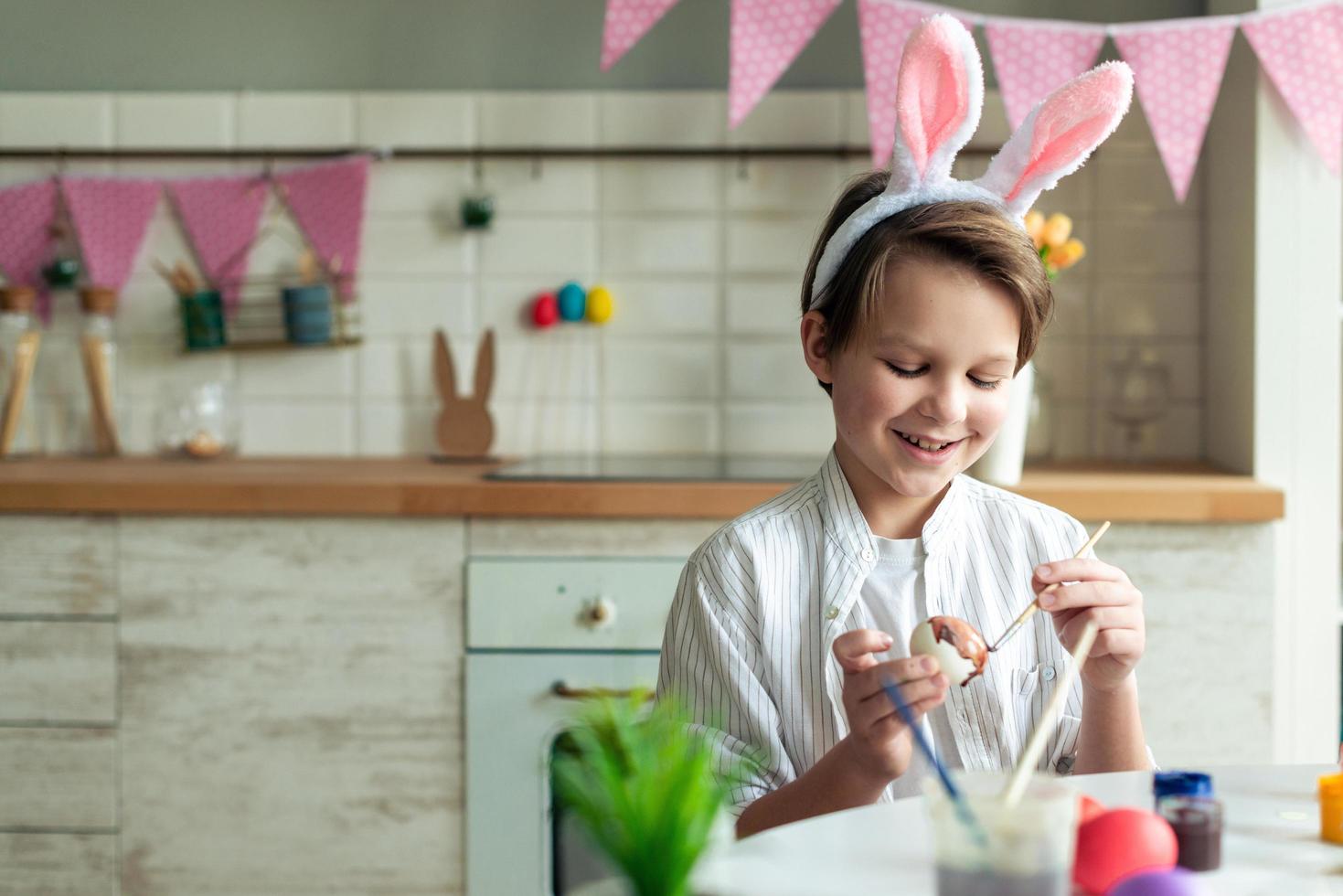 Close up of smiling boy sitting at table in kitchen and painting easter egg. photo