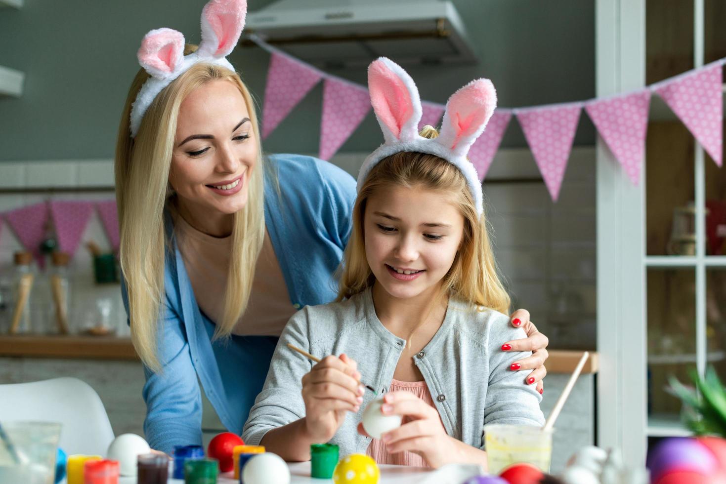 Happy Easter atmosphere. Funny, pretty mom making control, looking how her cute, small, joyful daughter draw, paint, decorate easter eggs, together wearing bunny ears photo