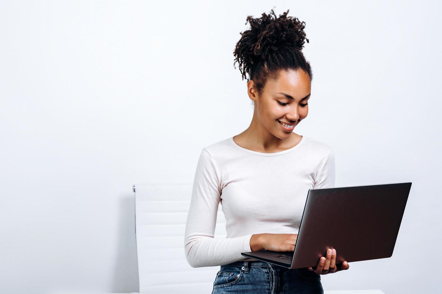 Foto de joven mujer de negocios alegre africana de pie sobre una pared blanca con ordenador portátil.