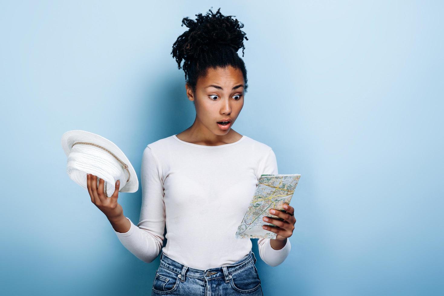 Shocked dark-skinned girl with a map and a hat in her hands isolated on a blue background photo