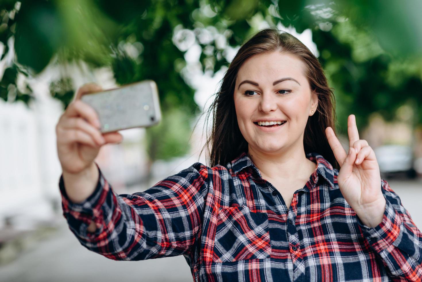 Smiling pretty girl showing peace gesture while taking a selfie isolated over city background photo