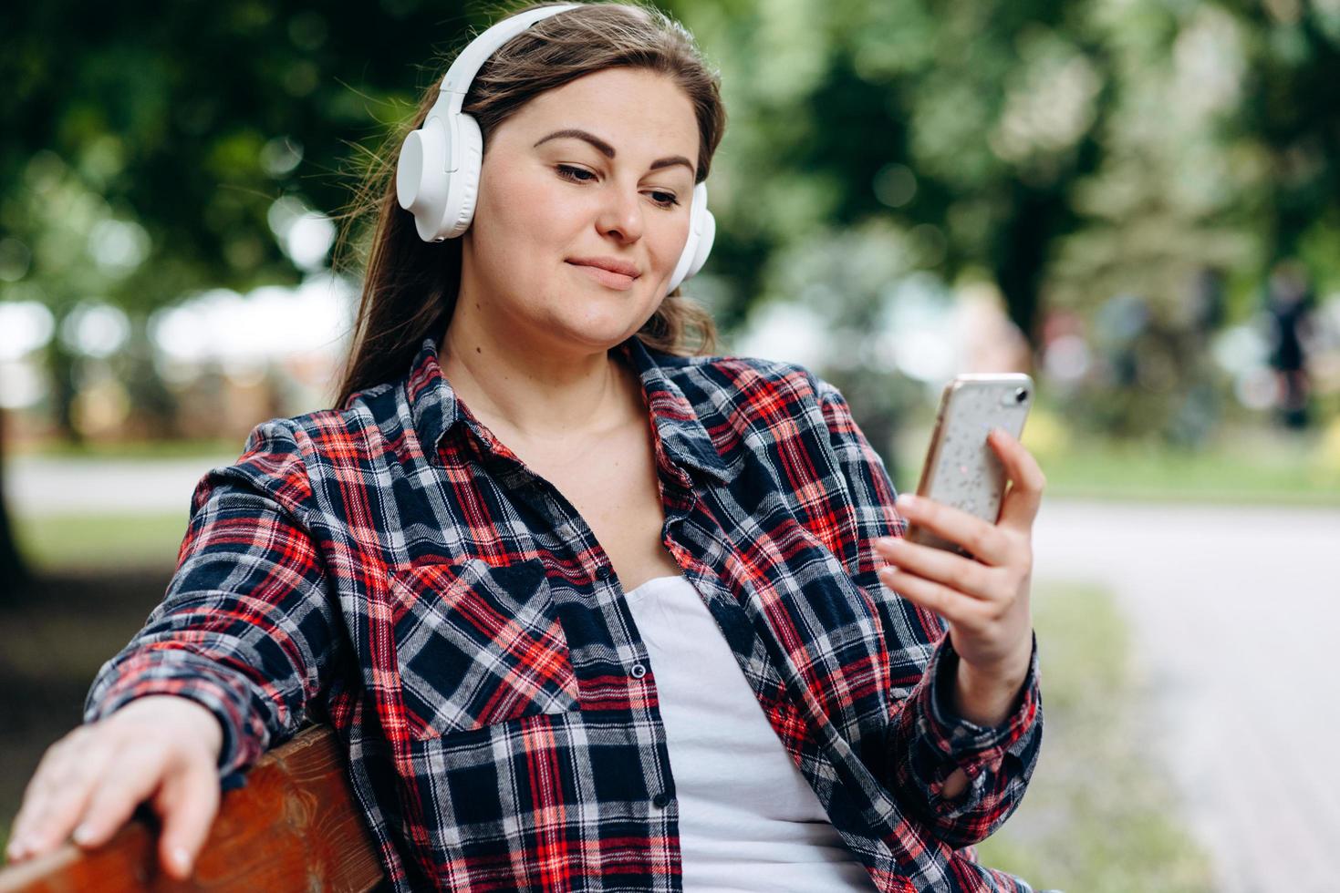 Cute woman, plus size, sitting on a bench in headphones, looking at a smartphone photo