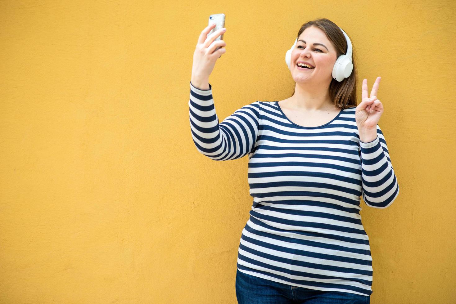 Con el telón de fondo de una pared naranja, una mujer alegre y sonriente muestra un gesto de paz y se toma una selfie en un teléfono inteligente foto