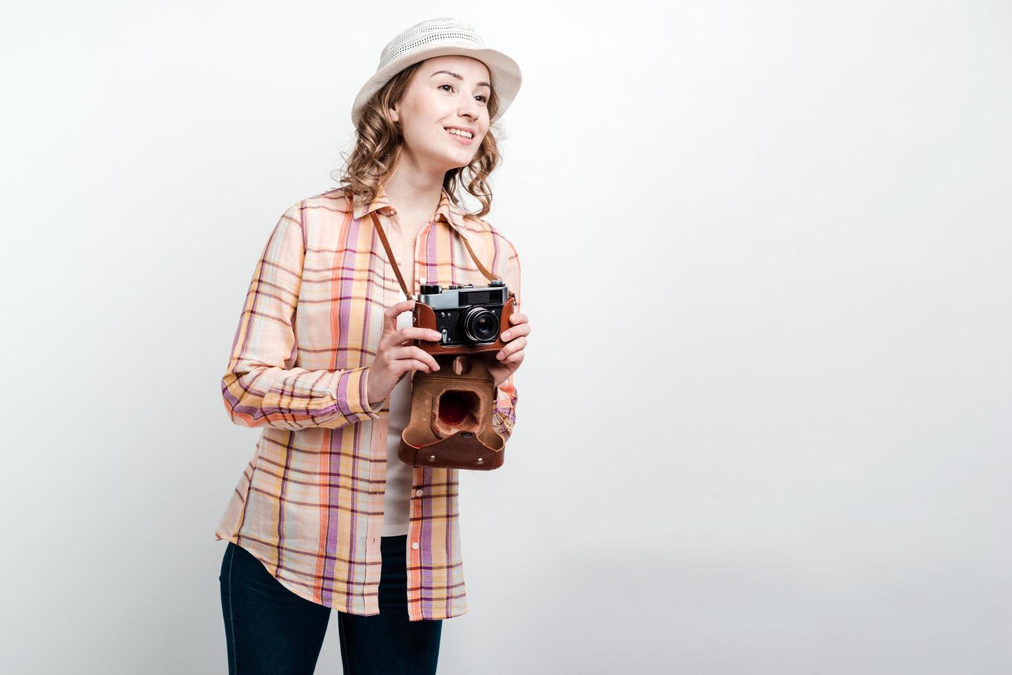 Chica con cámara retro y sombrero mira hacia otro lado sobre fondo de pared blanca foto