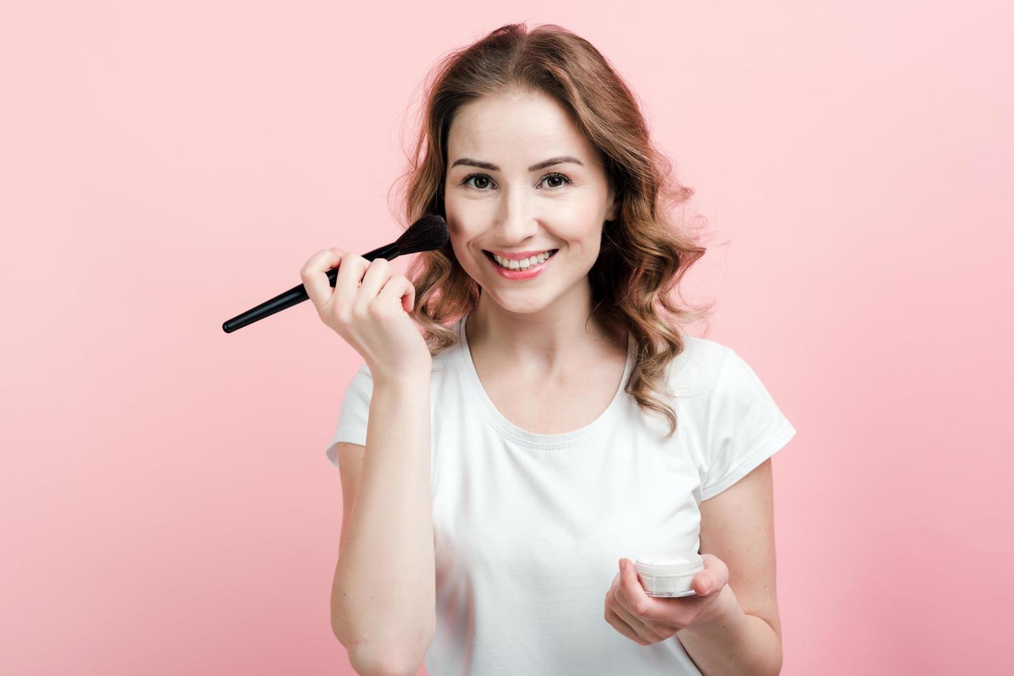 Linda y sonriente morena aplica maquillaje sobre fondo de pared rosa foto