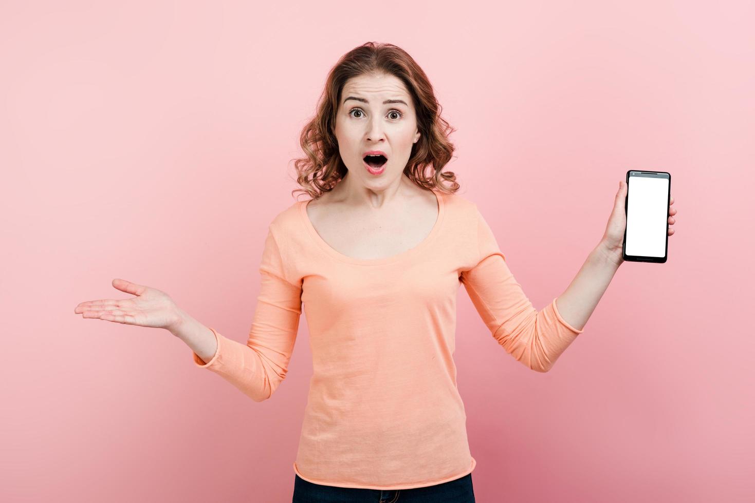 Impressed shocked dark haired girl surprised has smart phone isolated over pink studio wall. Copy space. photo