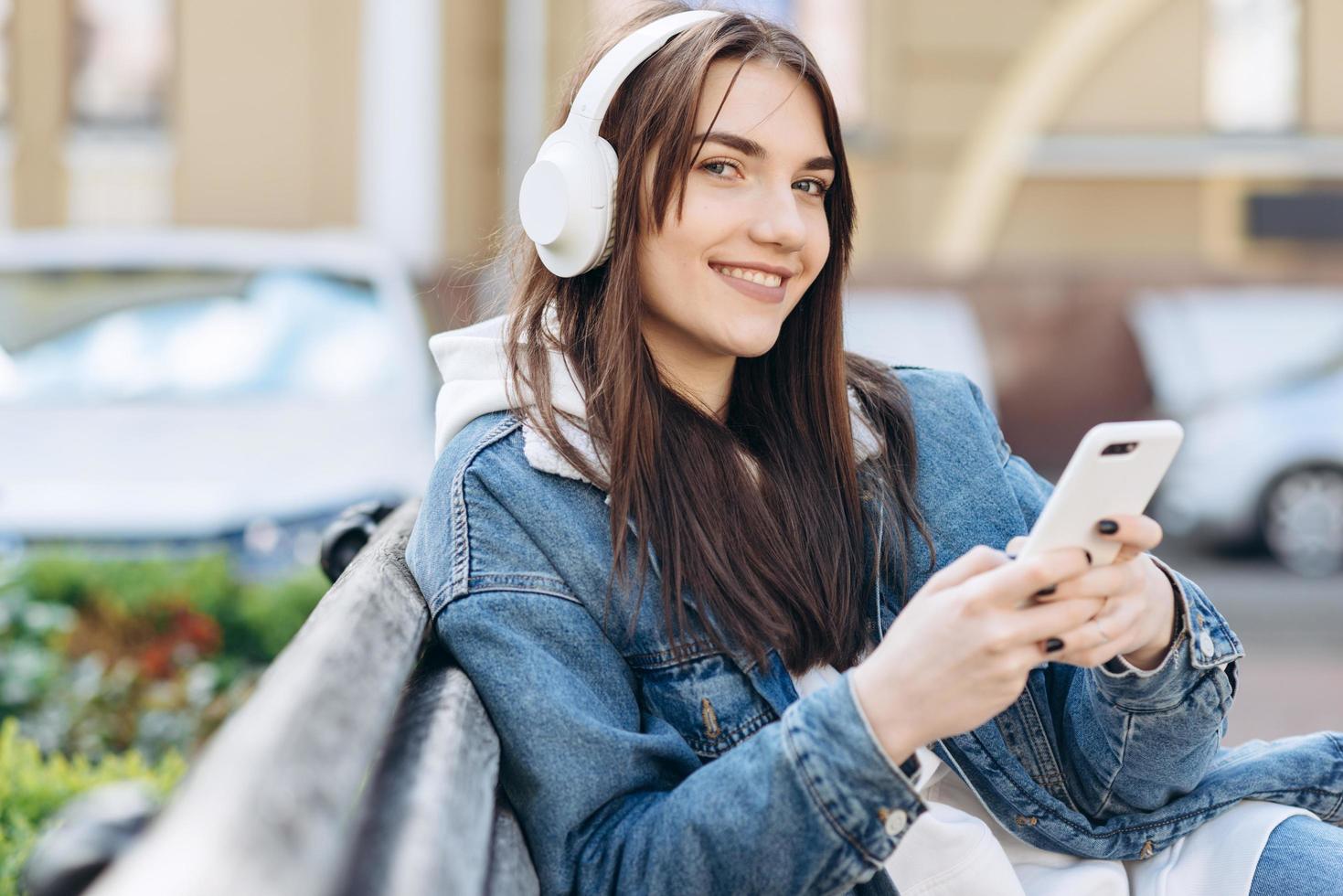 niña sonriente sentada en un banco y disfrutando, escuchando música en blanco, auriculares inalámbricos, sosteniendo un teléfono inteligente en la mano. calle con coches sobre un fondo borroso foto