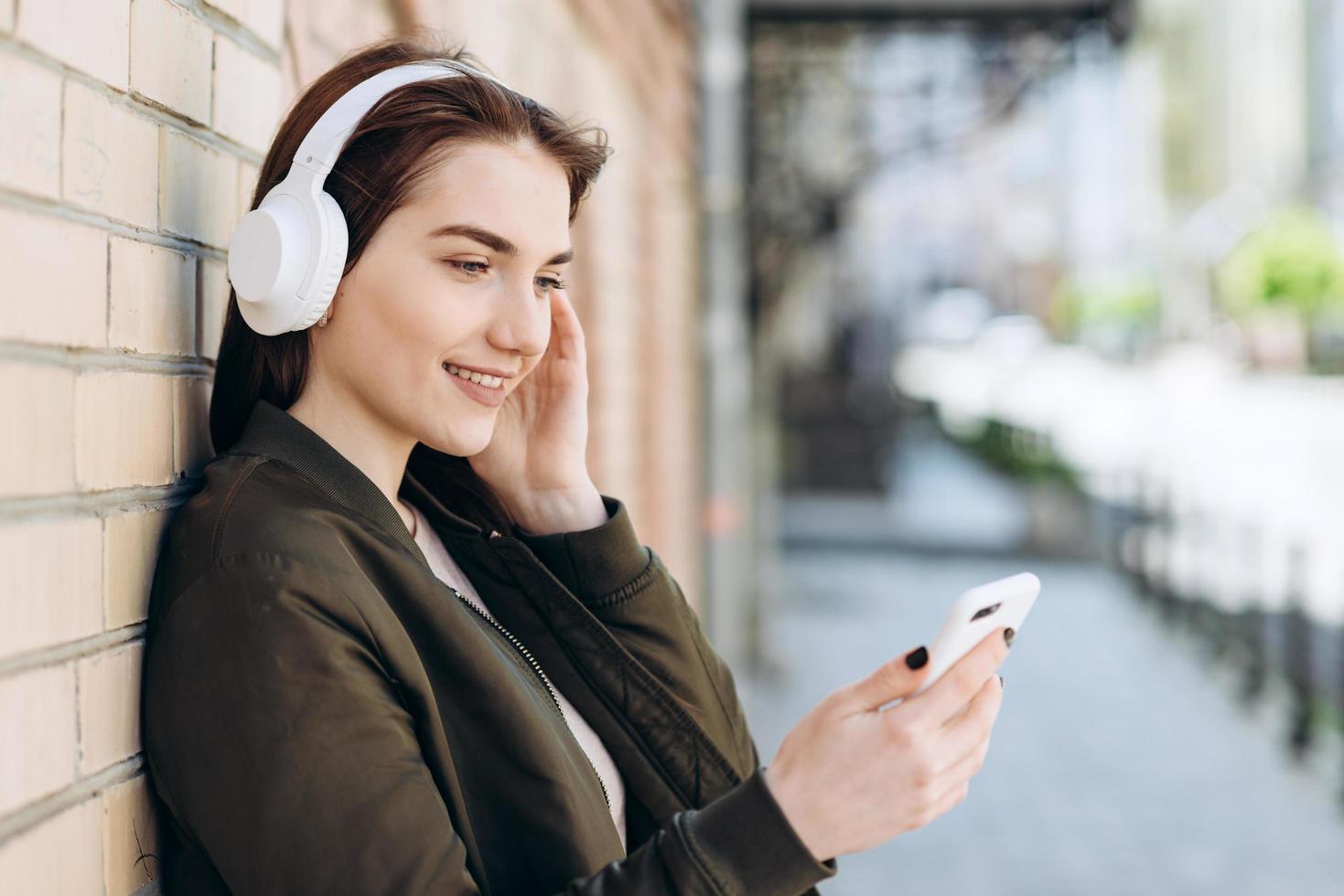 niña sonriente y alegre en auriculares inalámbricos se apoya contra una pared de ladrillos, incluye tu música favorita foto