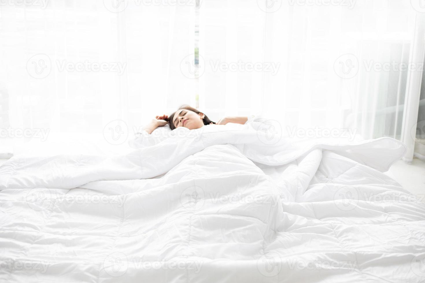 mujer asiática hermosa joven mujer sonriente sentada en la cama y estirándose por la mañana en el dormitorio después de despertarse en su cama completamente descansada y abrir las cortinas por la mañana para tomar aire fresco. foto
