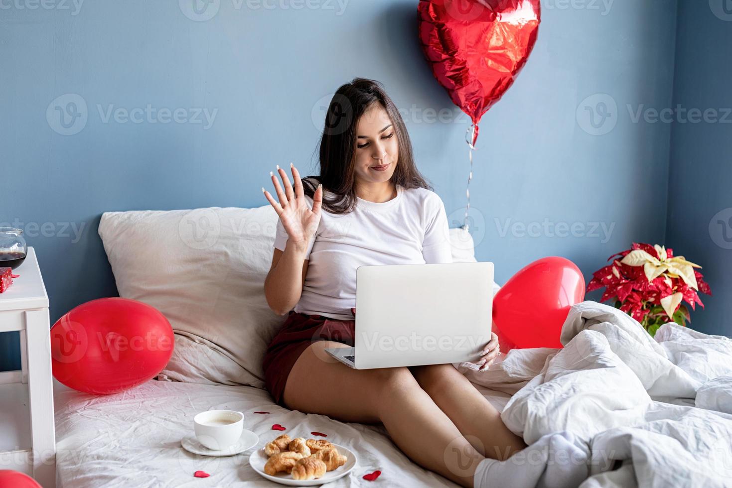 Feliz joven mujer morena sentada en la cama con globos en forma de corazón rojo trabajando en la computadora portátil foto