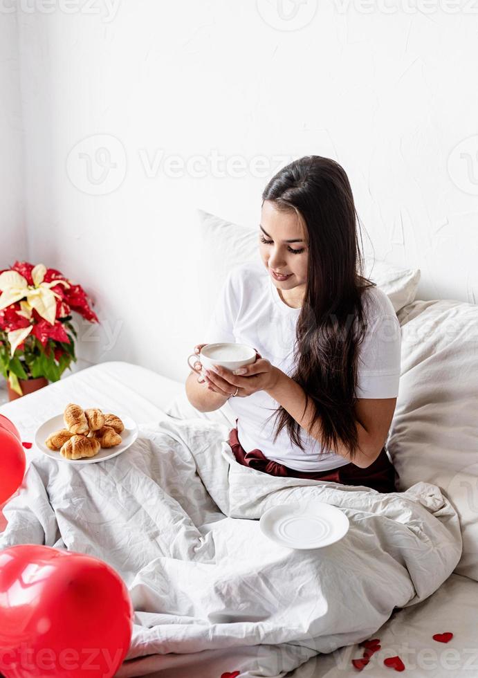 Joven mujer morena sentada despierta en la cama con globos en forma de corazón rojo y adornos bebiendo café comiendo croissants foto