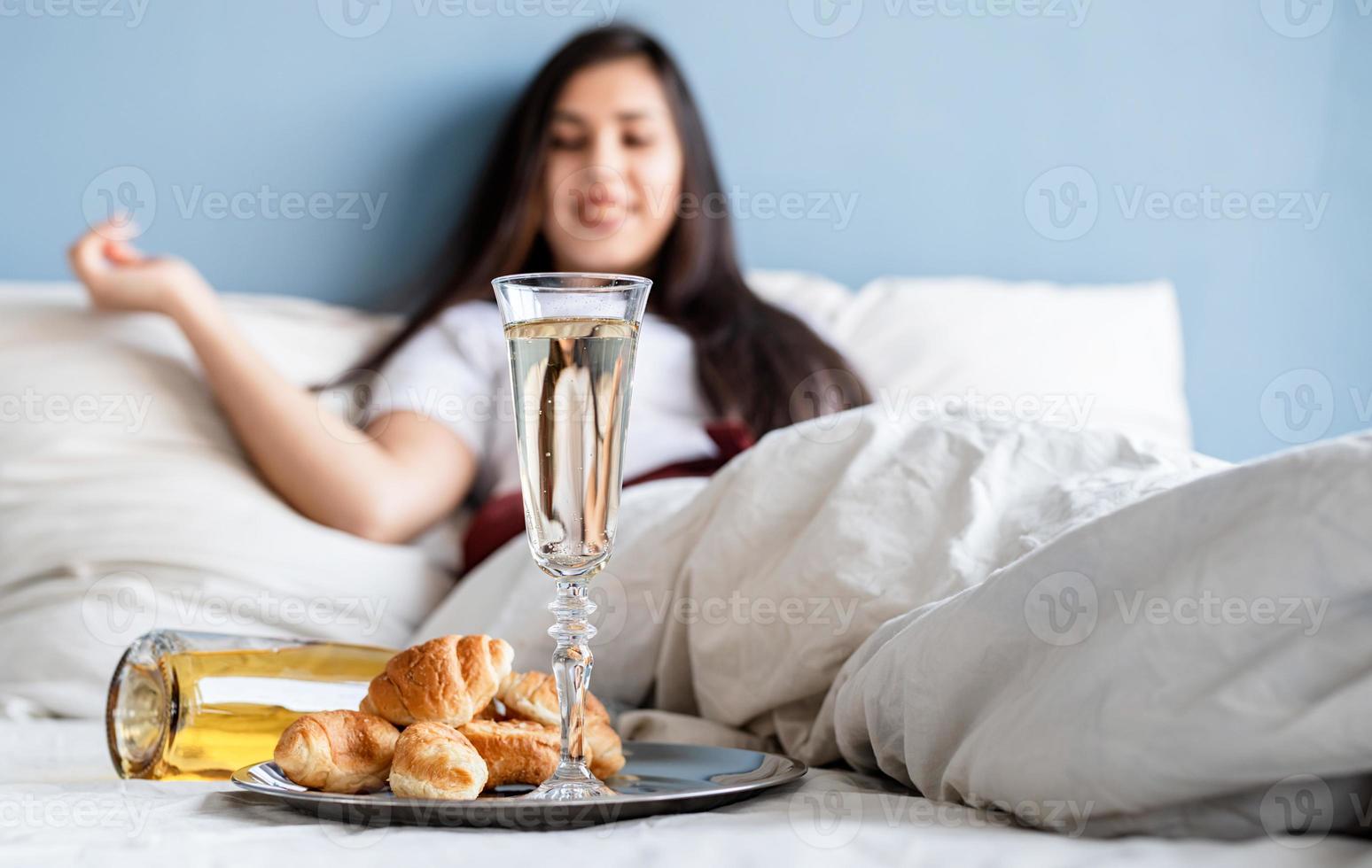 Joven mujer morena sentada despierta en la cama con globos en forma de corazón rojo y adornos bebiendo champán comiendo croissants foto