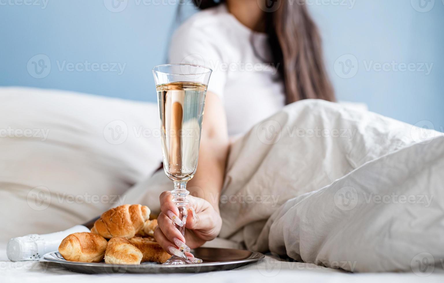 Young brunette woman sitting awake in the bed with red heart shaped balloons and decorations drinking champagne eating croissants photo