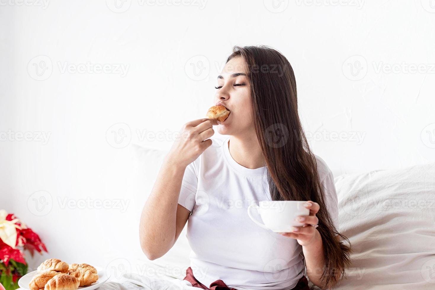 Mujer joven morena sentada en la cama con croissants comiendo foto