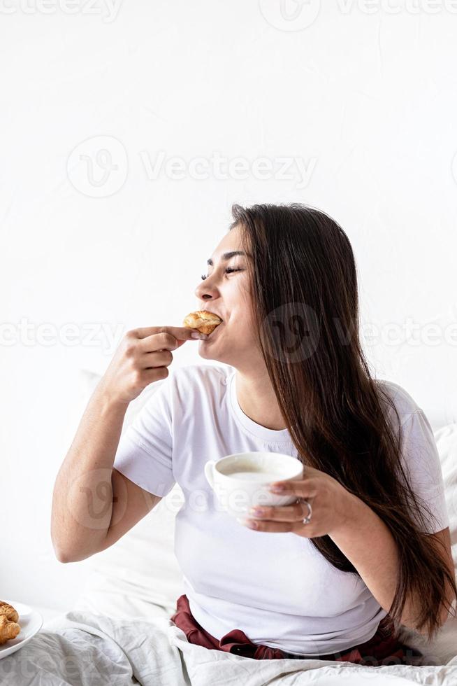 Mujer joven morena sentada en la cama con croissants comiendo foto