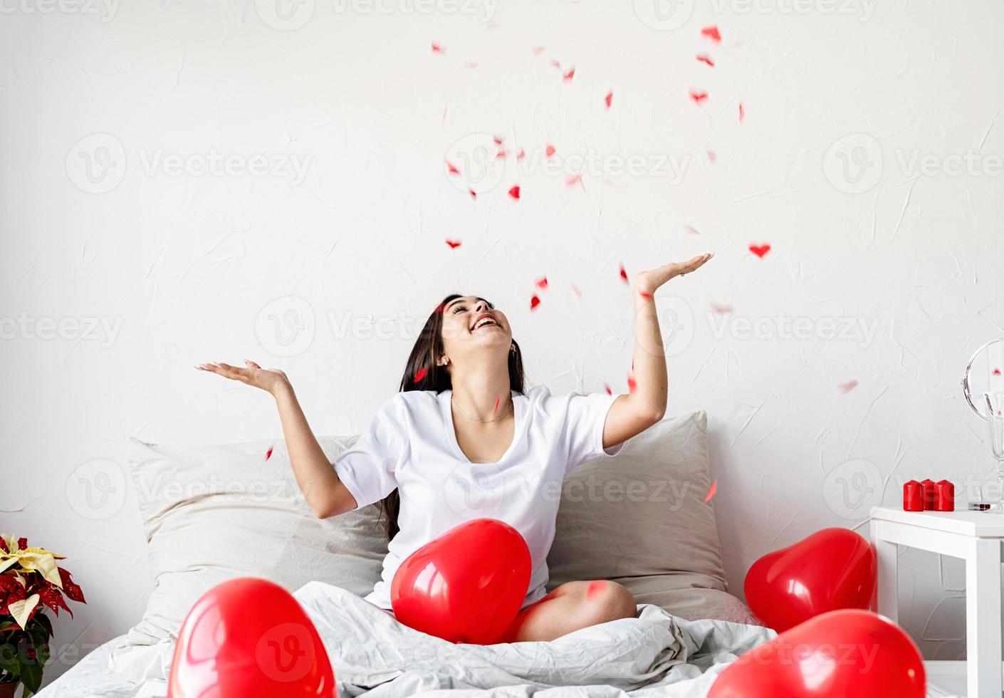 Feliz joven mujer morena sentada en la cama con globos en forma de corazón rojo lanzando confeti en el aire foto