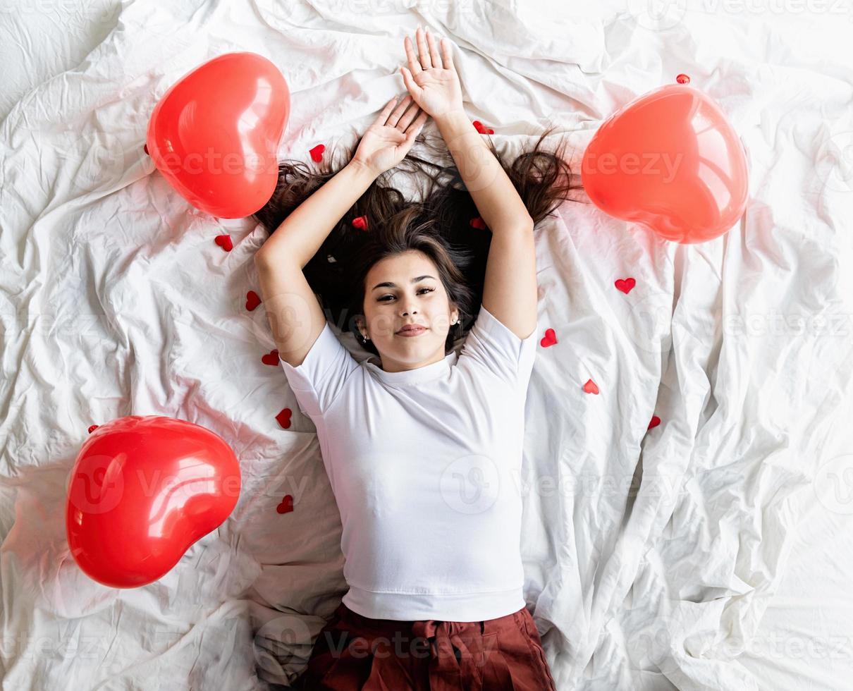 Joven mujer morena feliz acostado en la cama con globos y decoraciones en forma de corazón rojo foto