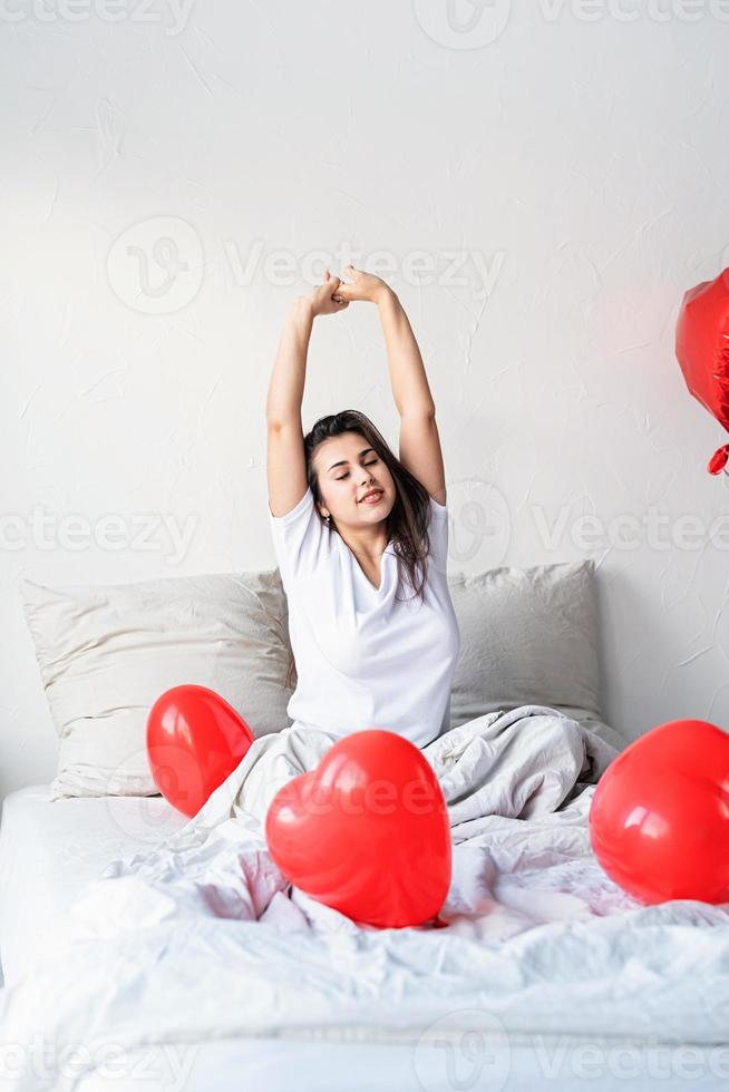 Feliz joven mujer morena sentada despierta en la cama con globos en forma de corazón rojo foto