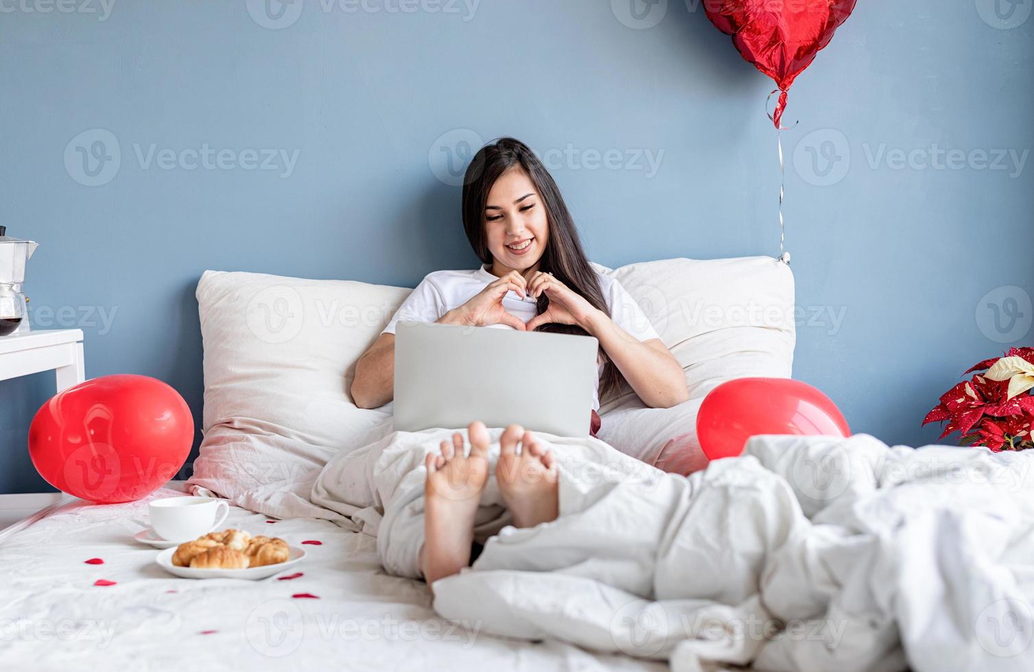 Joven mujer morena feliz sentada en la cama con globos en forma de corazón rojo charlando con su novio en la computadora portátil que muestra el gesto del corazón con las manos foto