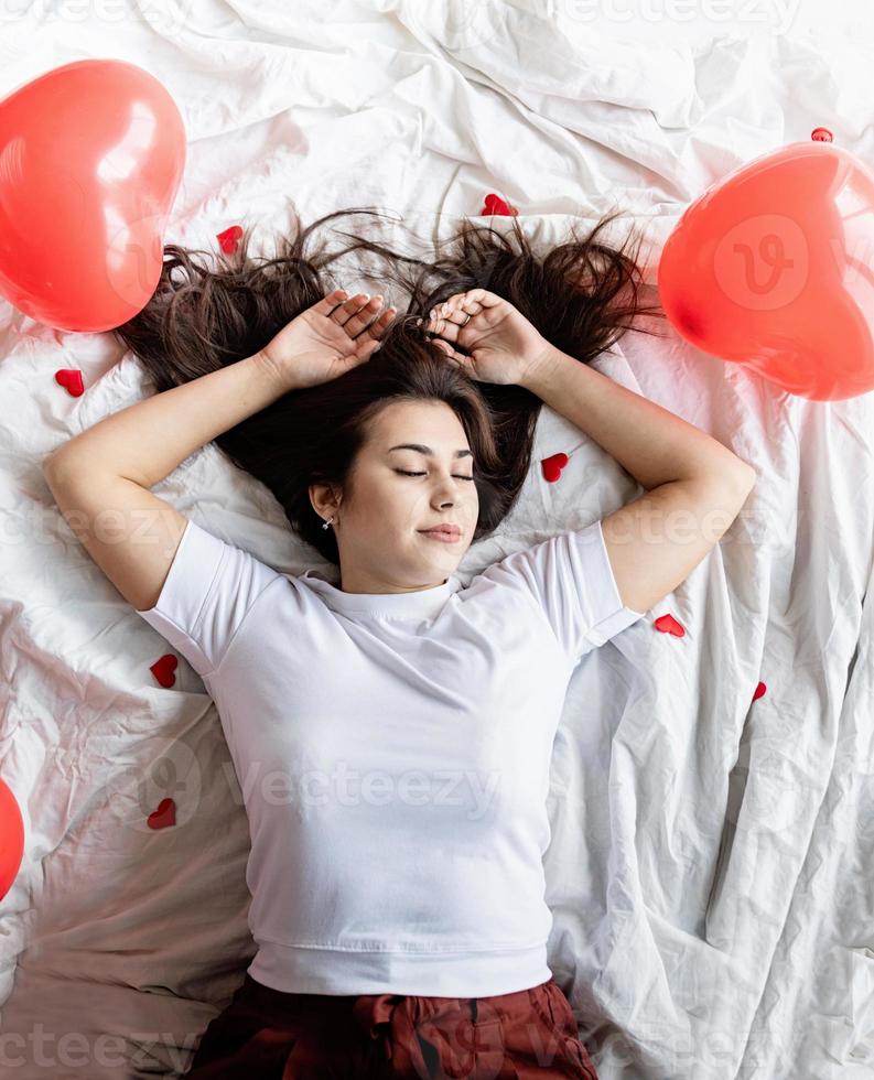 Young happy brunette woman laying in the bed with red heart shaped balloons and decorations photo