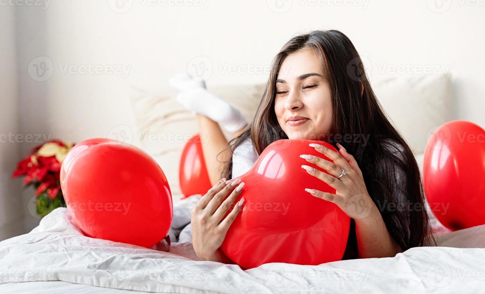 Young happy brunette woman laying in the bed with red heart shaped balloons photo