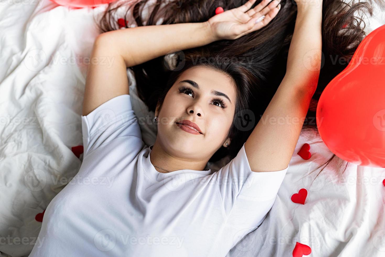 Joven mujer morena feliz acostado en la cama con globos y decoraciones en forma de corazón rojo foto