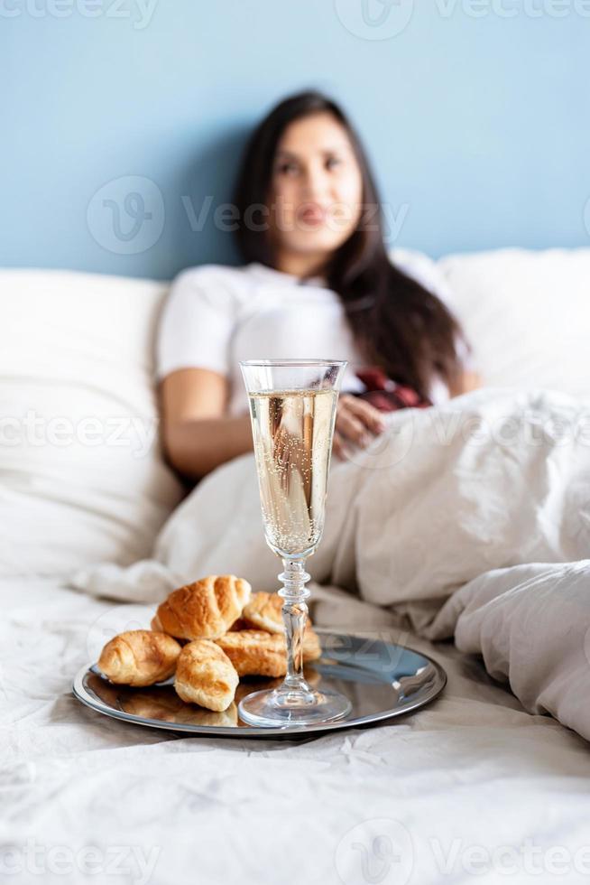 Joven mujer morena sentada despierta en la cama con globos en forma de corazón rojo y adornos bebiendo champán comiendo croissants foto