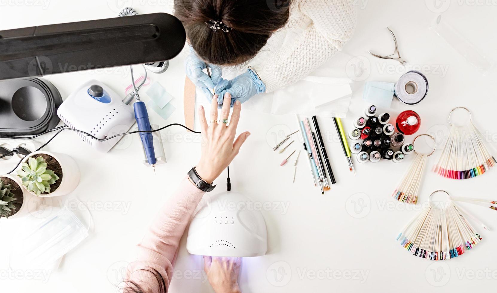 Maestro de manicura en máscara y guantes poniéndose esmalte de gel en las uñas de un cliente vista superior foto