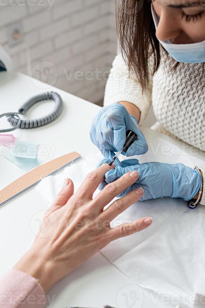 Maestro de manicura en máscara y guantes poniéndose esmalte de gel en las uñas de un cliente foto