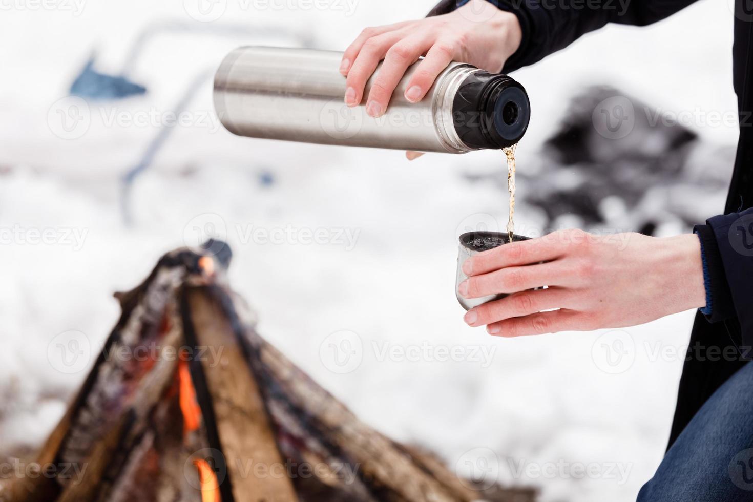 primer plano de hombre de manos de viajero vierte té de frascos en una taza en el bosque cerca de la hoguera. foto