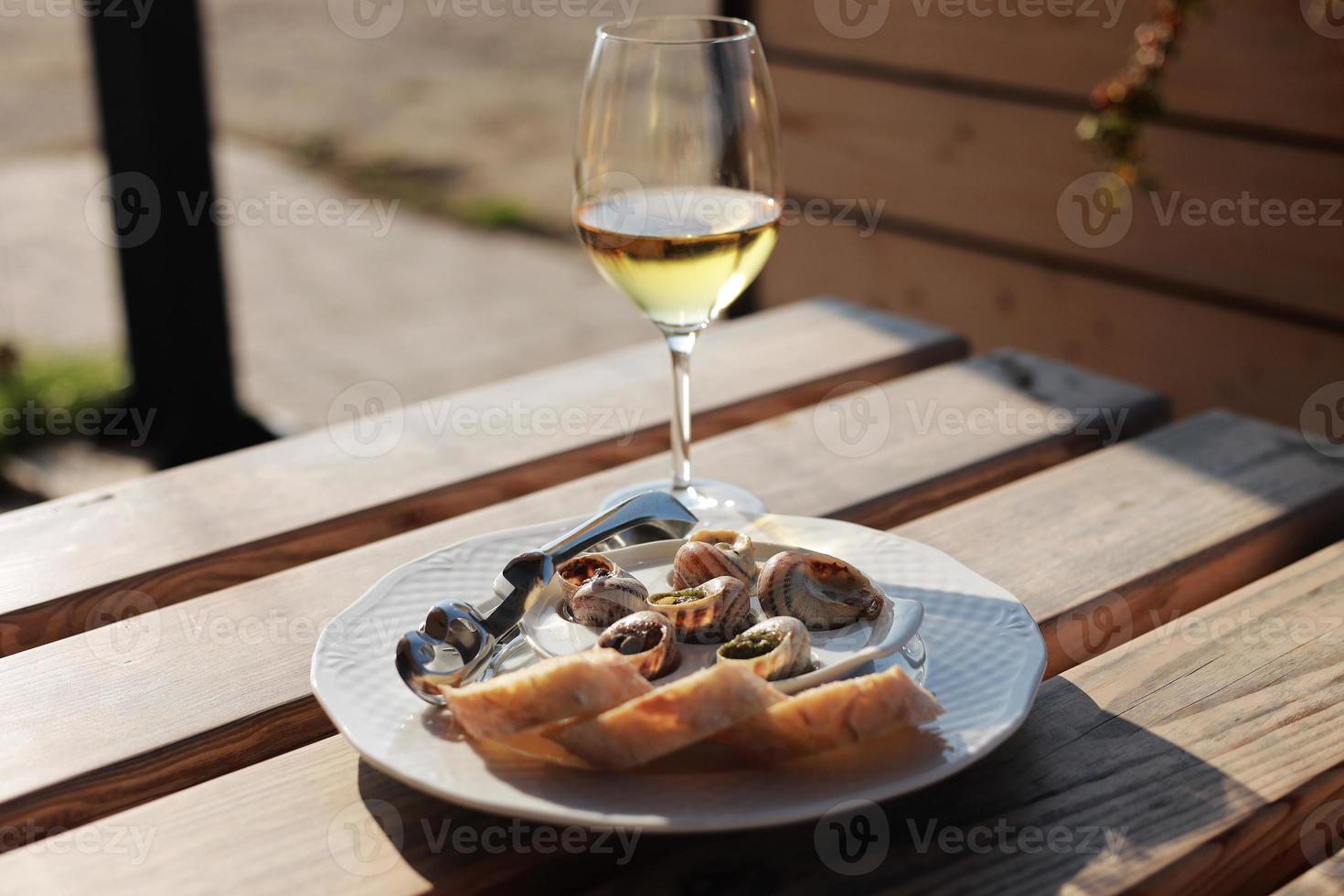 Glass with wine and dish with the snail shells, served on a white classic plate, with various sauces, pesto, truffle, cheese, pieces of French baguette, fork for a shell, wooden table in outdoor cafe photo